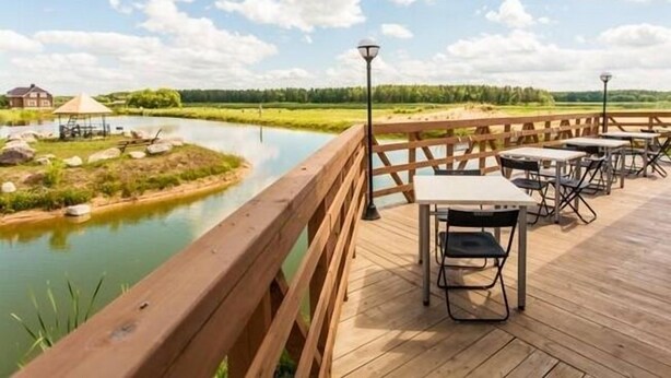 Una terraza de madera con mesas y sillas que da a un río tranquilo, rodeada de vegetación y un cielo despejado.
