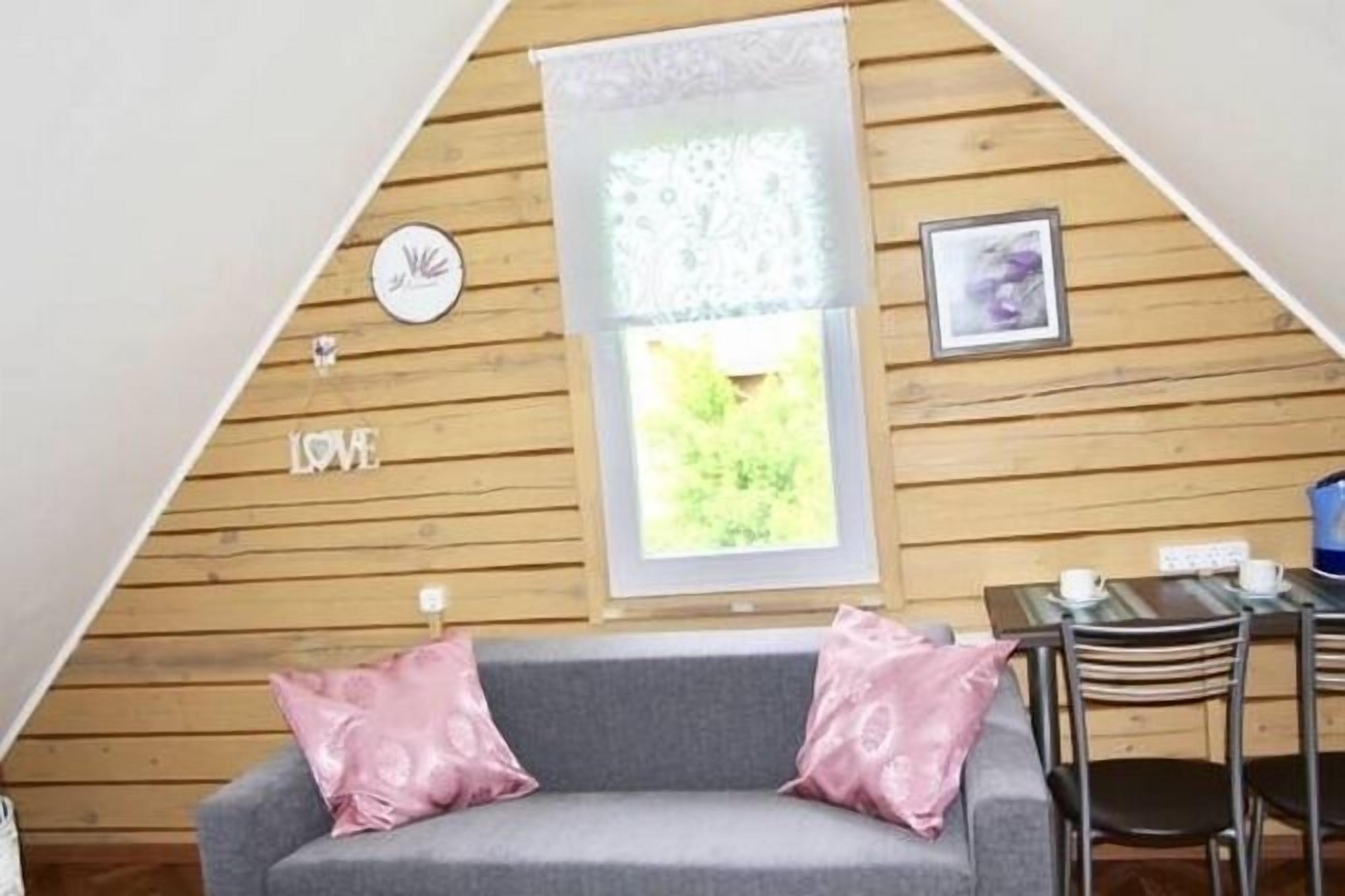 A cozy room with a sloped ceiling, wooden walls, a grey sofa with pink cushions, a small table with chairs, and a window with a view of greenery.