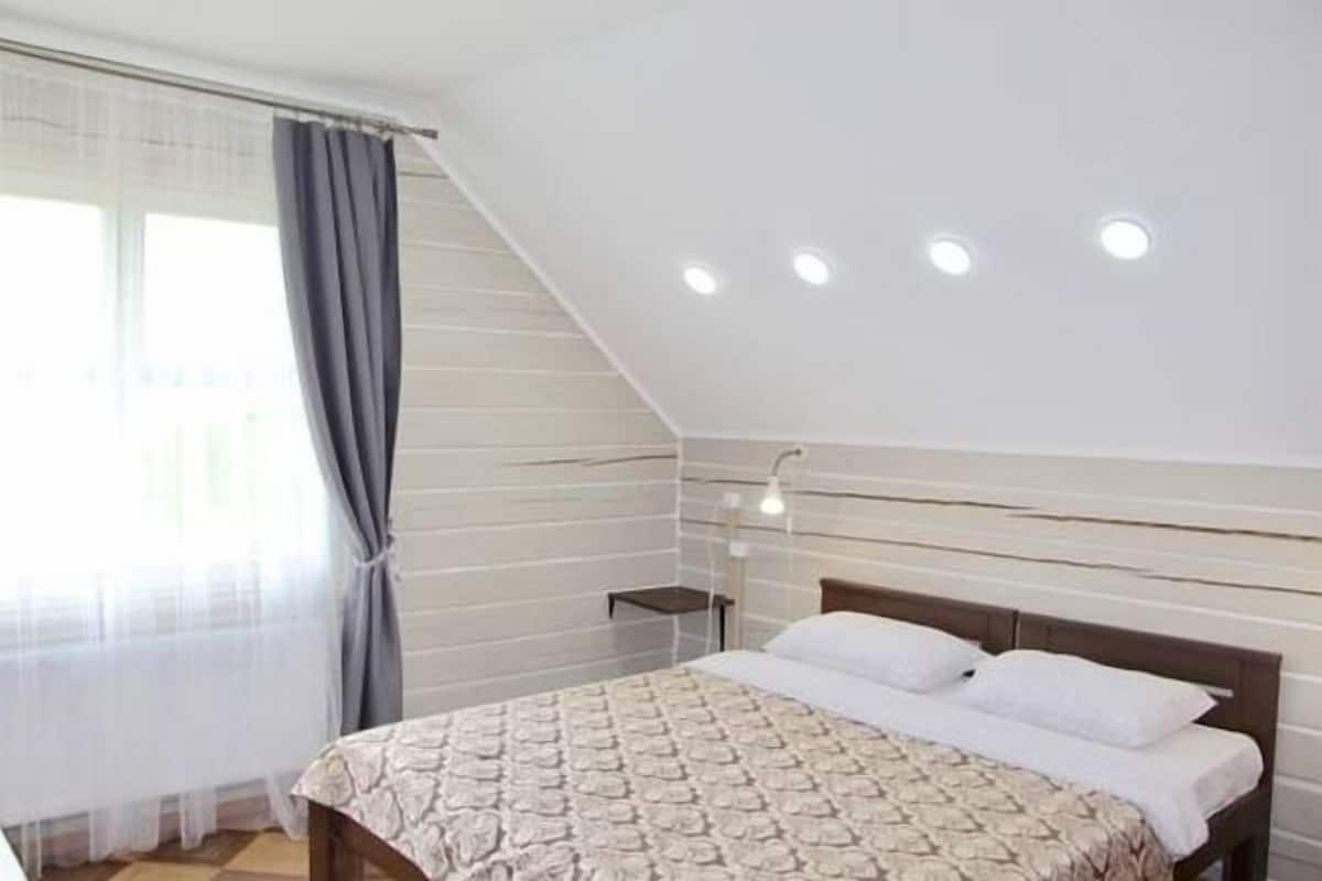 A bedroom with a sloped ceiling, a bed with a patterned bedspread, a bedside table, and a wall-mounted lamp.