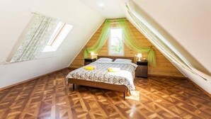 Un dormitorio con techo inclinado, piso de madera, cama con colcha estampada y ventana con cortinas.