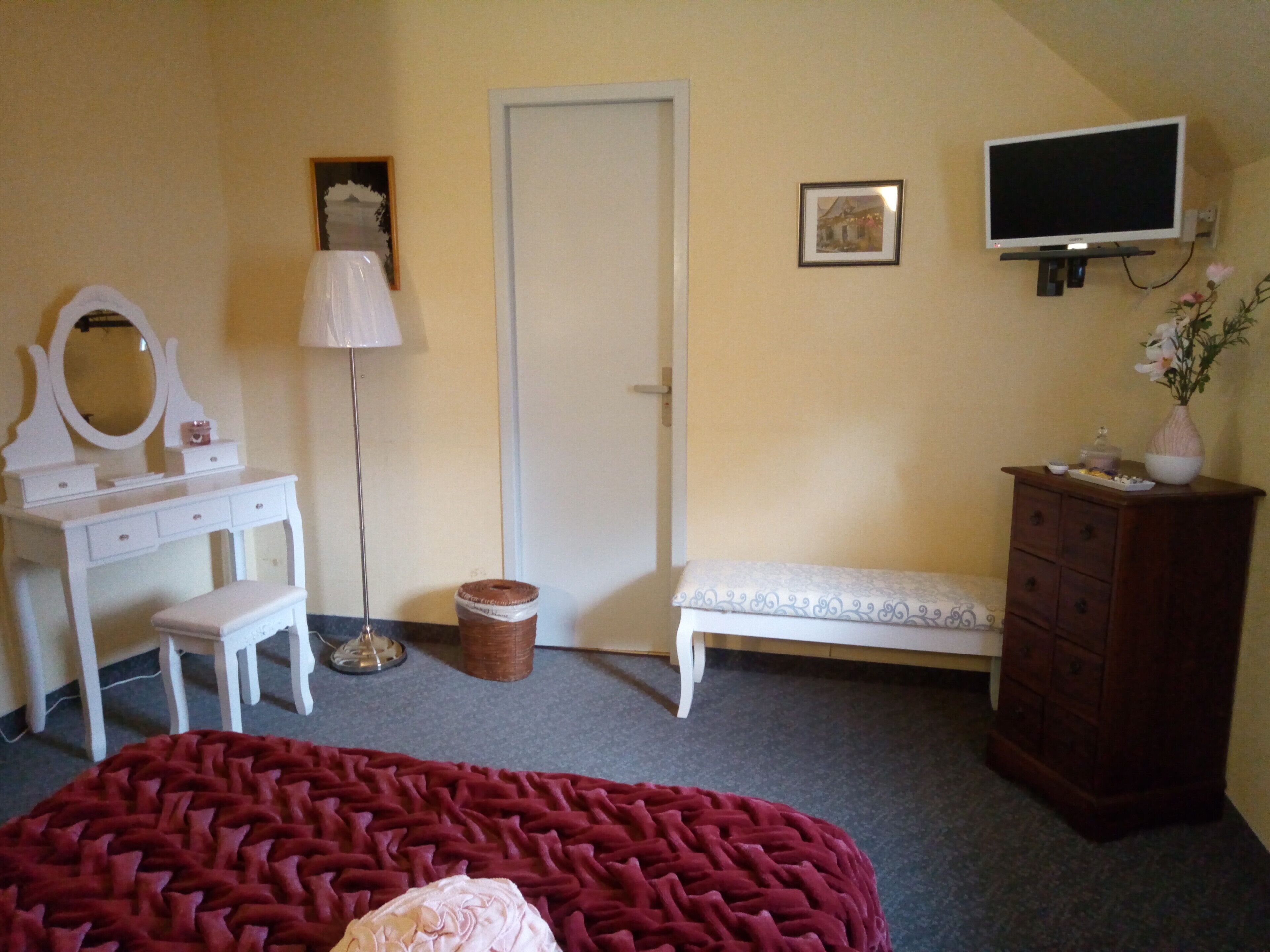 A bedroom with a bed, a dresser, a vanity with a mirror, a lamp, a door, and a television on the wall.