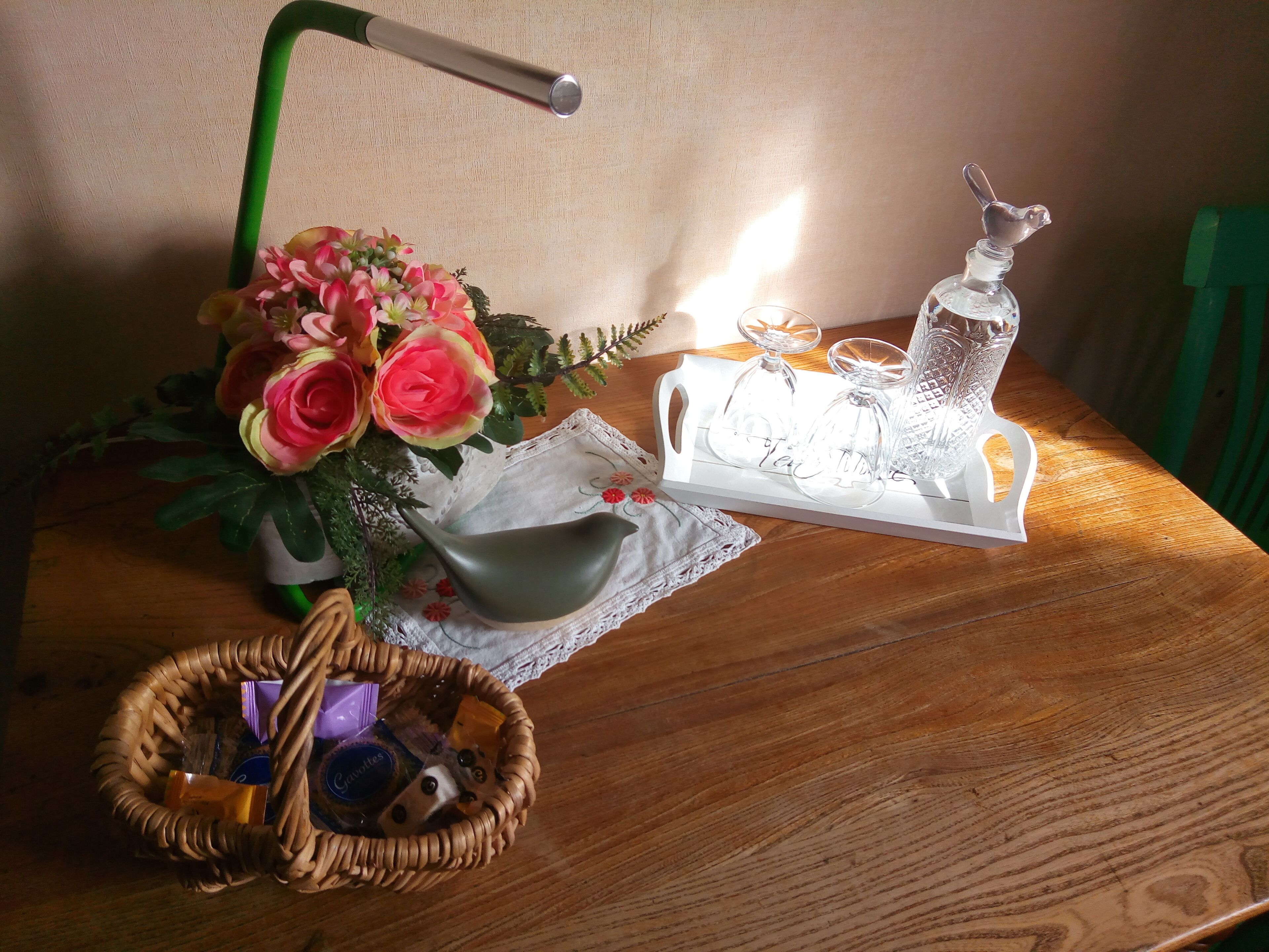 Une table en bois, sur laquelle se trouvent un panier en osier, un vase de fleurs, une lampe à manche verte et un plateau avec de la verrerie et un objet décoratif.