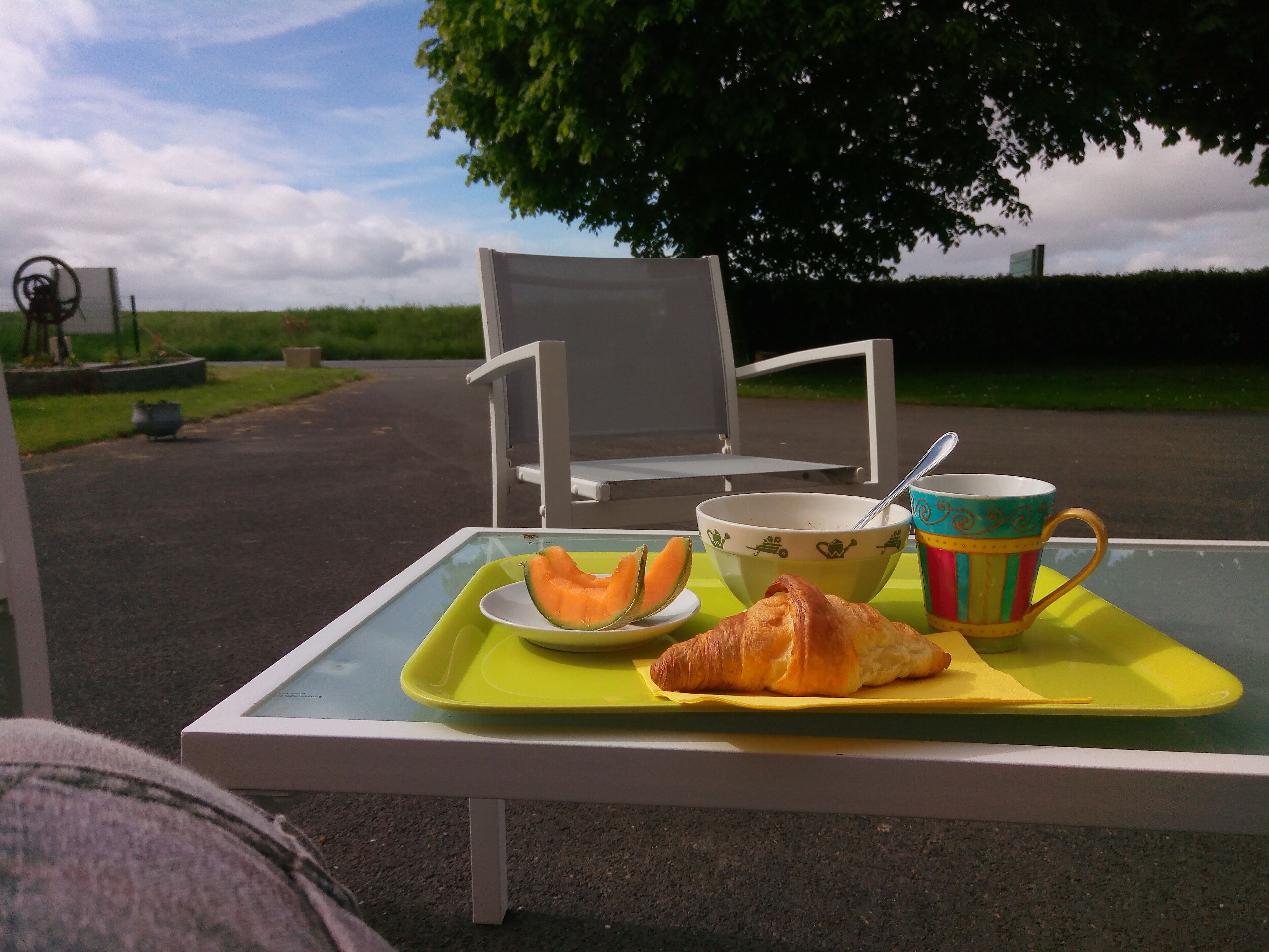 A table with a croissant, melon, bowl, and mug, set outdoors with a chair and a tree in the background.