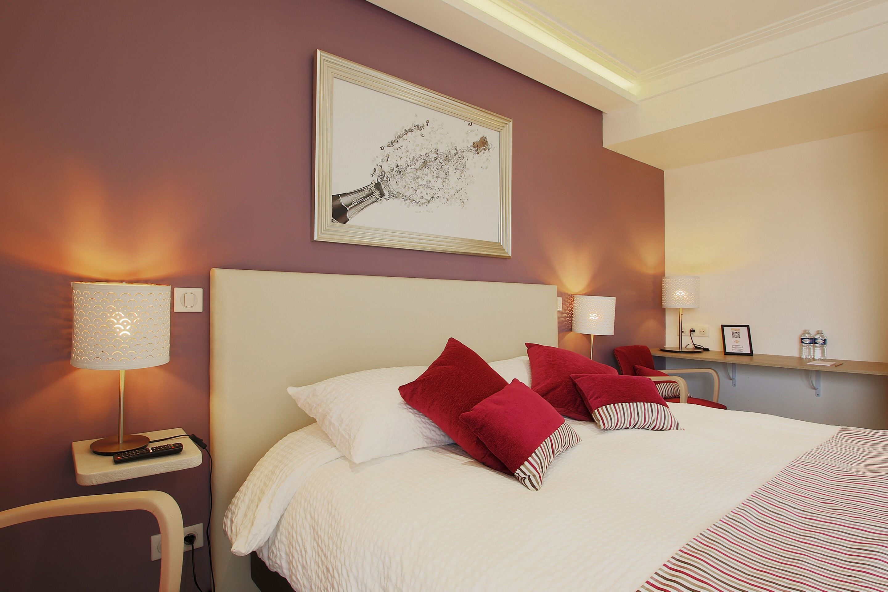 A hotel room with a large bed, red and white pillows, a framed picture on the wall, and bedside lamps.