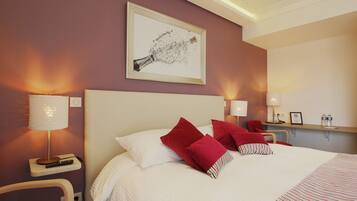A hotel room with a large bed, red and white pillows, a framed picture on the wall, and bedside lamps.