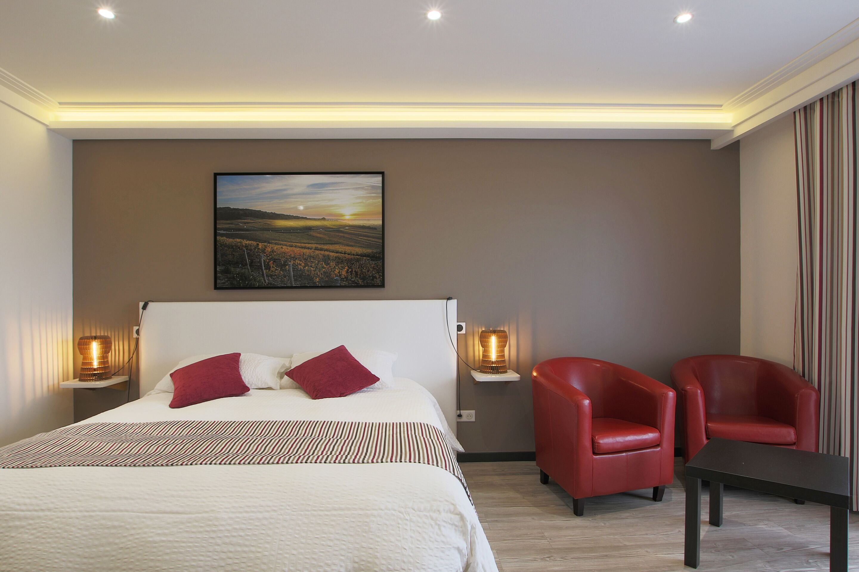 Habitación de hotel con una cama grande, dos sillas rojas, una mesita y un cuadro en la pared.