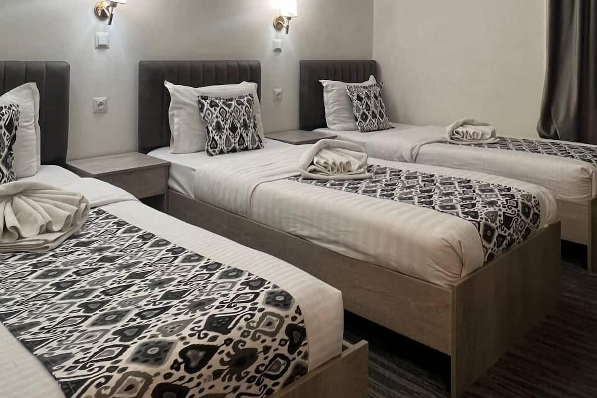 A hotel room with four single beds, each with a patterned bedspread, two wall-mounted light fixtures, and a window with curtains.