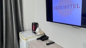 A hotel room with a television displaying 'ORZU HOTEL' on the screen, a small desk with a phone and remote controls, a coffee maker, and a mini-fridge. - Orzu Hotel (Tashkent)