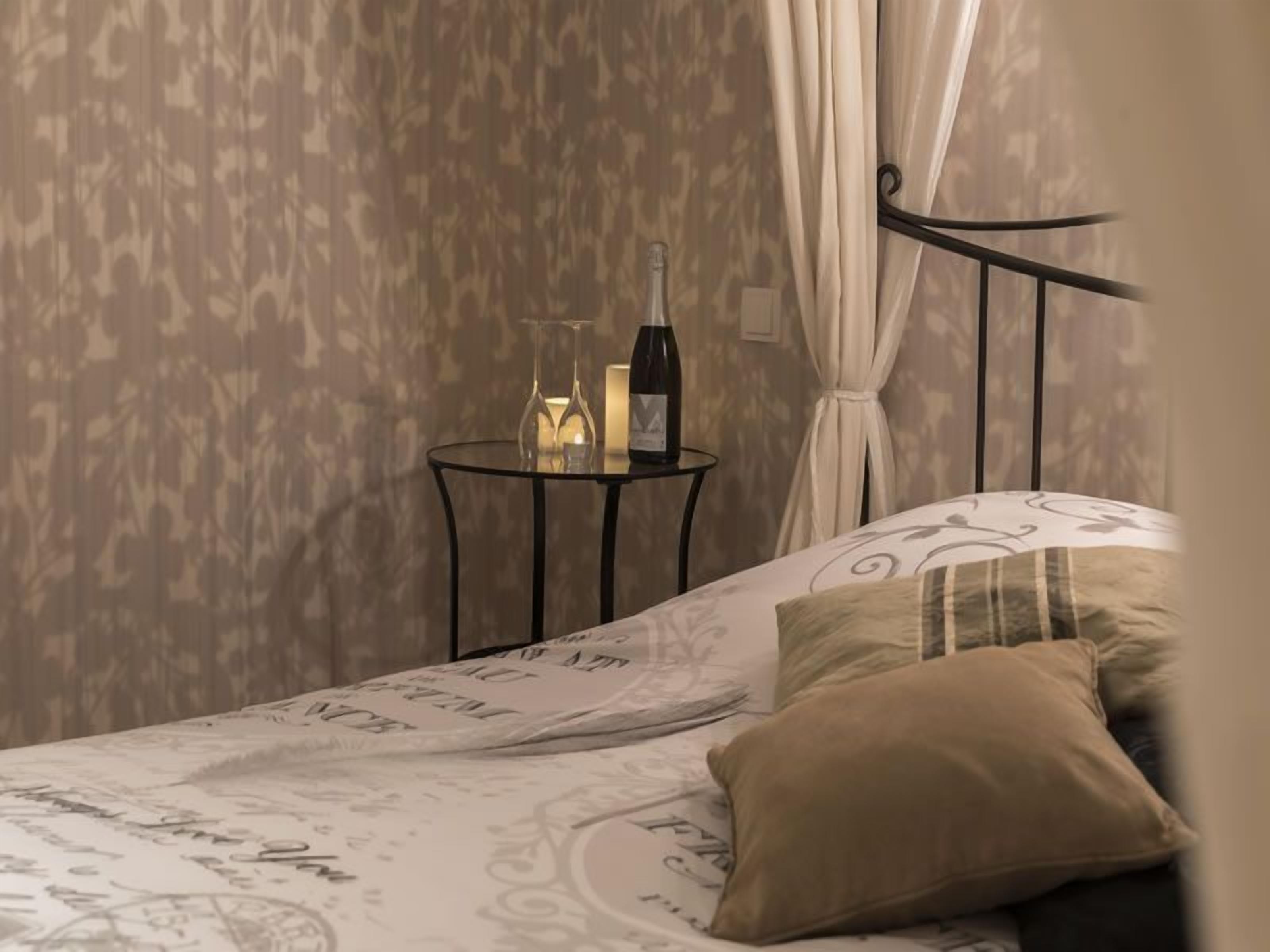 A bedroom with a bed, a bedside table with a bottle and glasses, and a curtain.