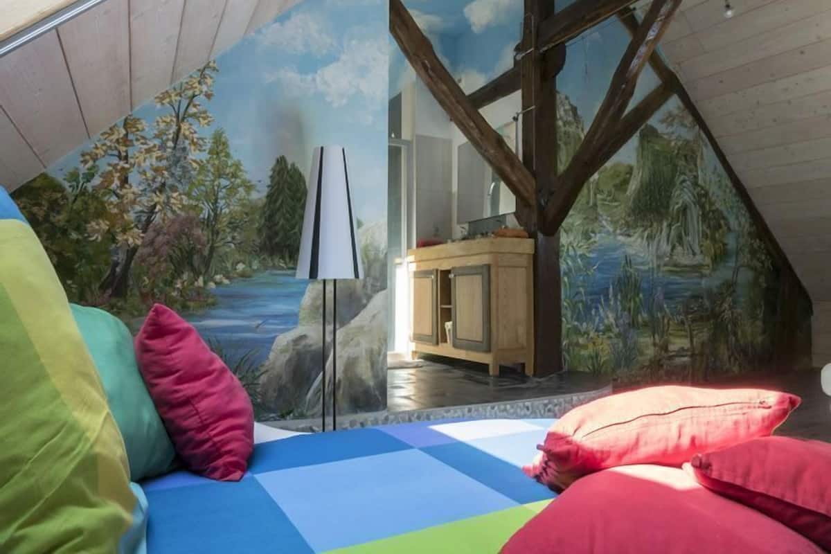 A cozy room with a sloped ceiling, a mural of a forest scene, a bed with colorful bedding, and a wooden cabinet.