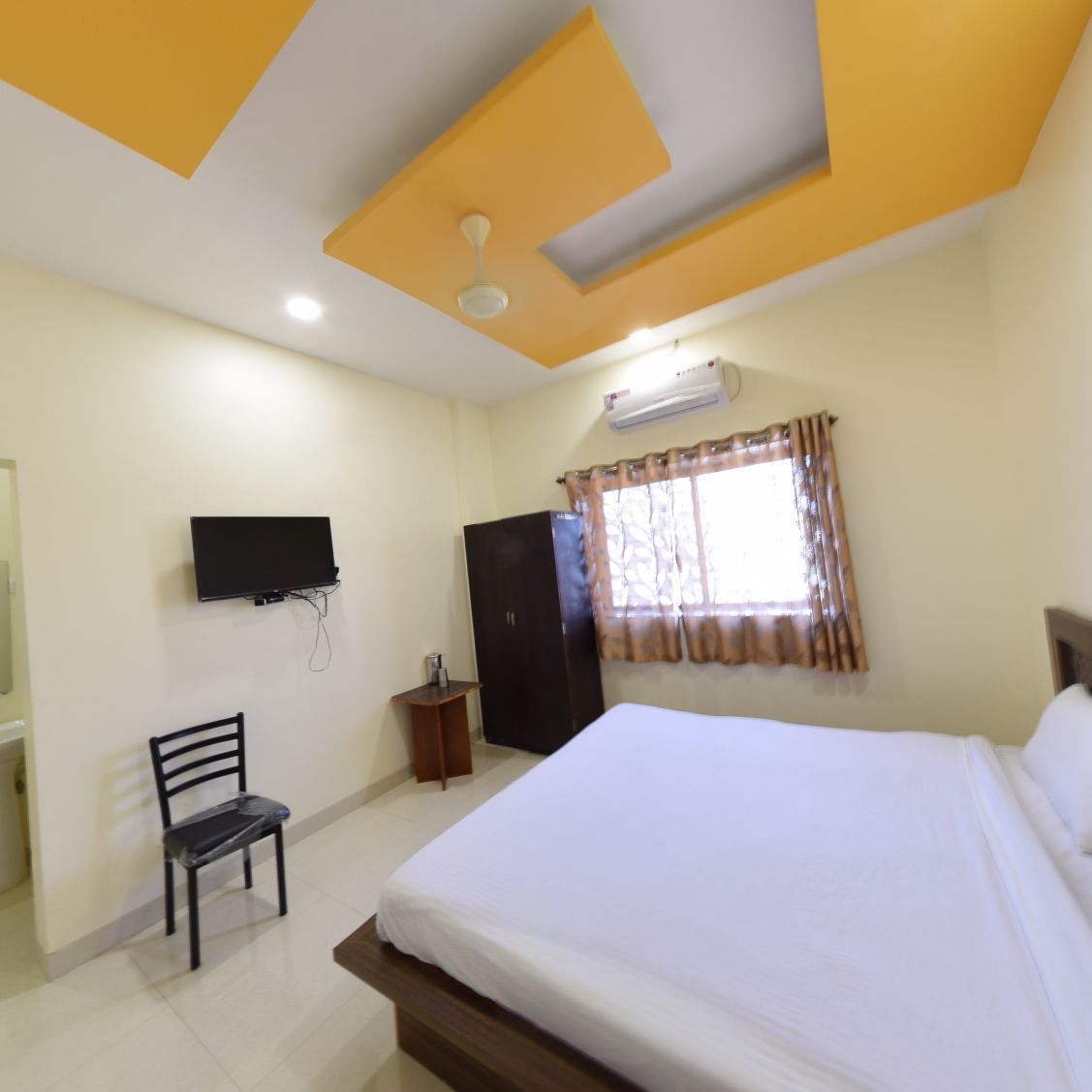 A hotel room with a bed, a TV mounted on the wall, a ceiling fan, and a window with curtains.