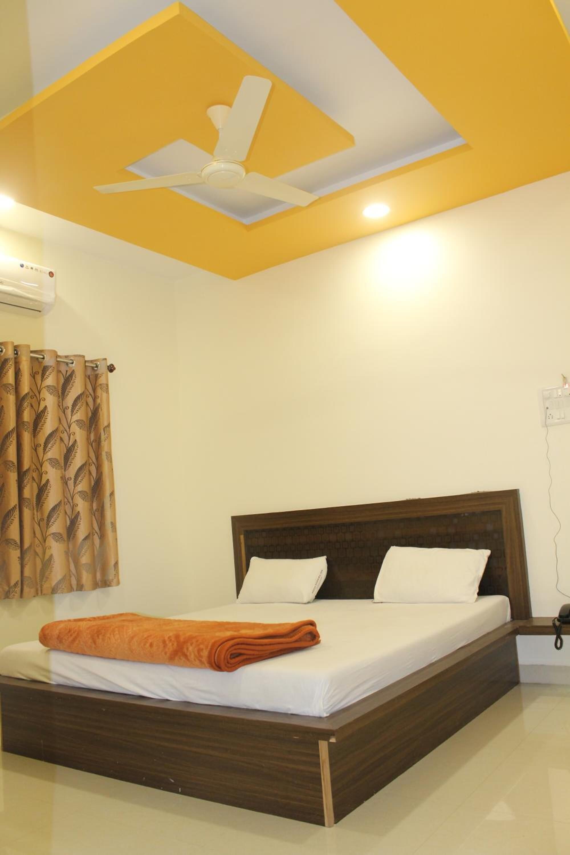 Una cama bien hecha con sábanas blancas y una manta naranja, cabecera de madera y ventilador de techo en una habitación con detalles en amarillo y techo blanco.