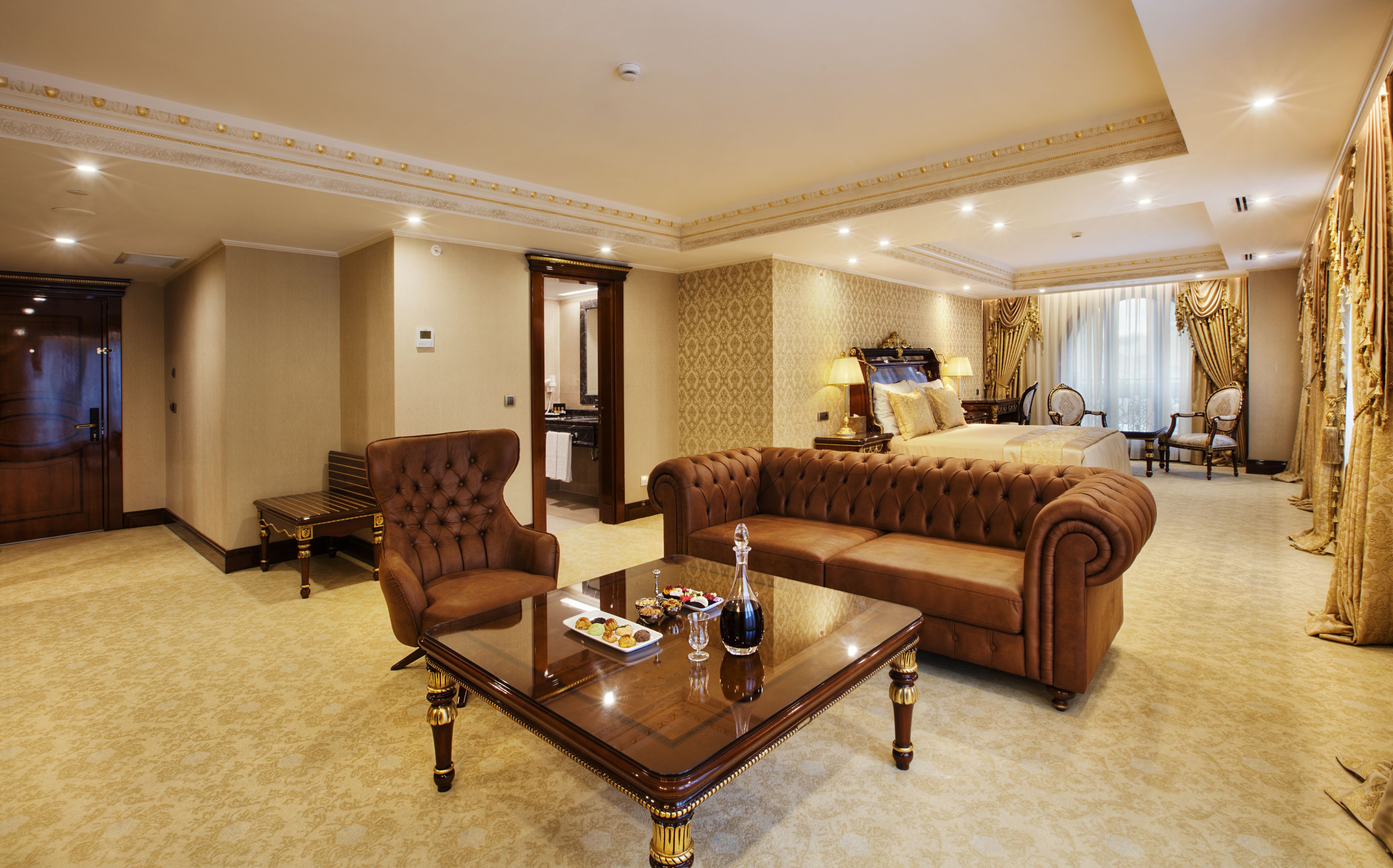 A spacious hotel room with a large bed, a sitting area with a sofa and armchairs, a dining area with a table and chairs, and a separate bathroom.