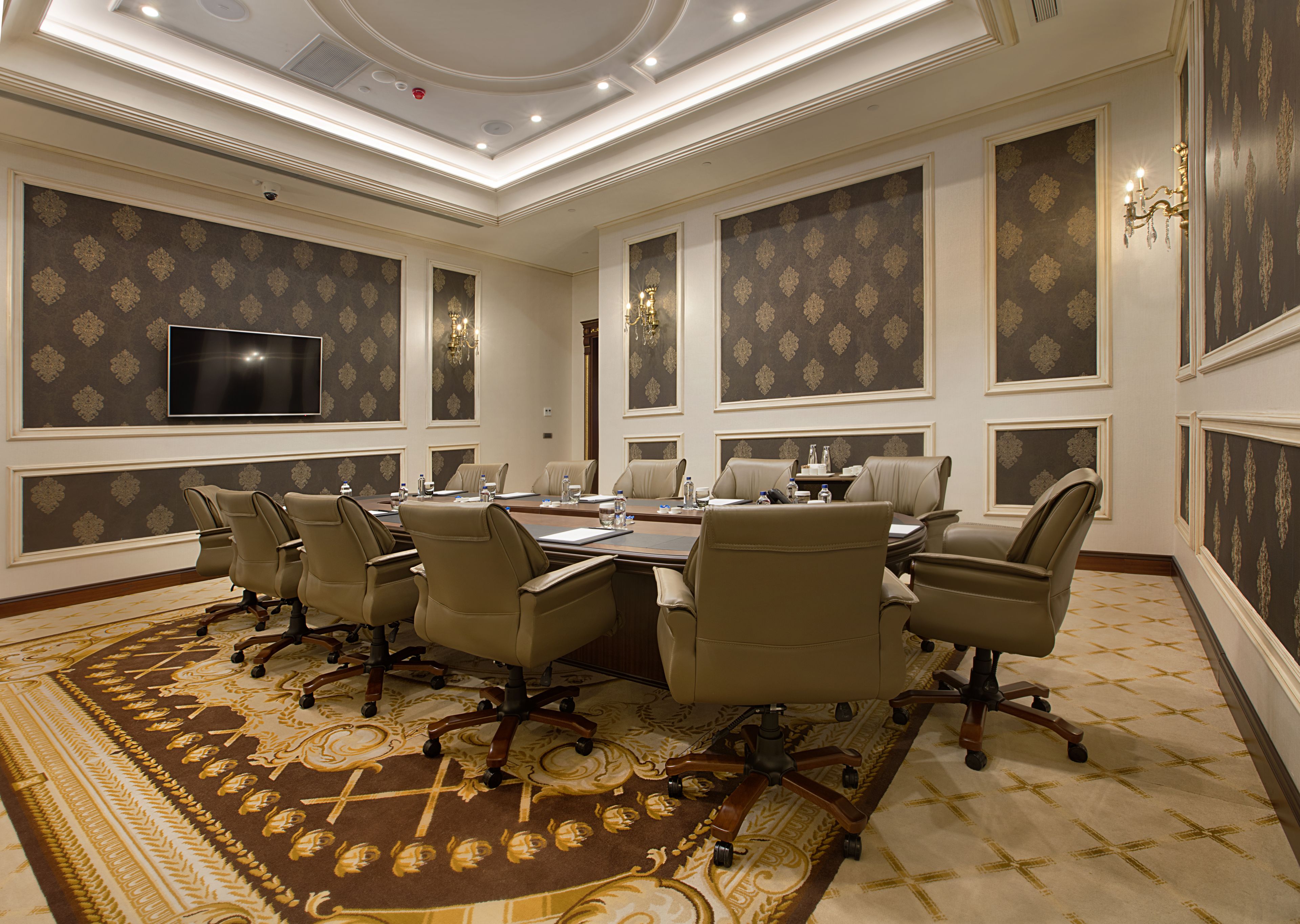 A conference room with a large oval table, multiple chairs, a flat-screen TV, and patterned wallpaper.