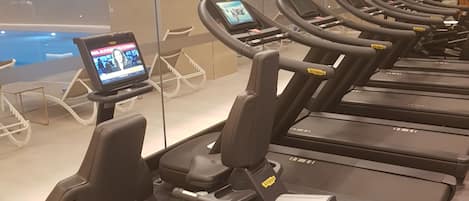 Una hilera de bicicletas estáticas con pantallas digitales, un muro de cristal y una piscina al fondo.