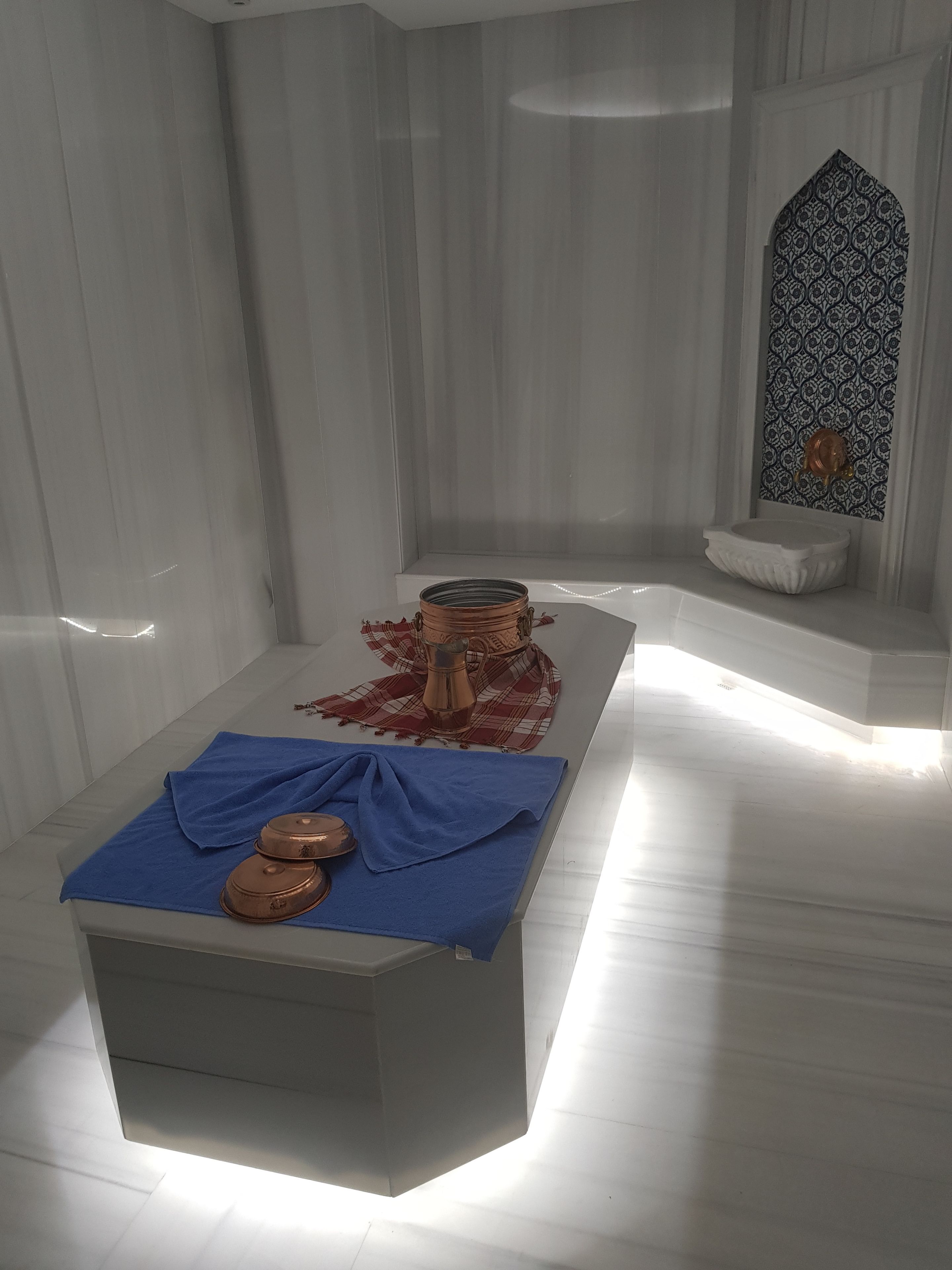 Una habitación con una mesa blanca, un mantel azul y ollas de cobre. La pared tiene un diseño y una ventana pequeña con un adorno.