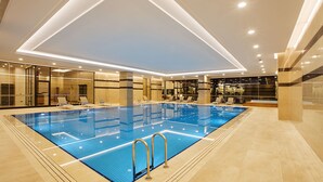 Une piscine intérieure moderne, avec une eau d’un bleu limpide, entourée de chaises longues et dotée d’un plafond bien éclairé.