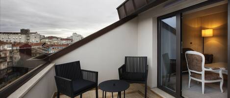 Uma varanda com duas cadeiras pretas e uma mesa redonda pequena, com vista para um horizonte urbano com edifícios e uma ponte.
