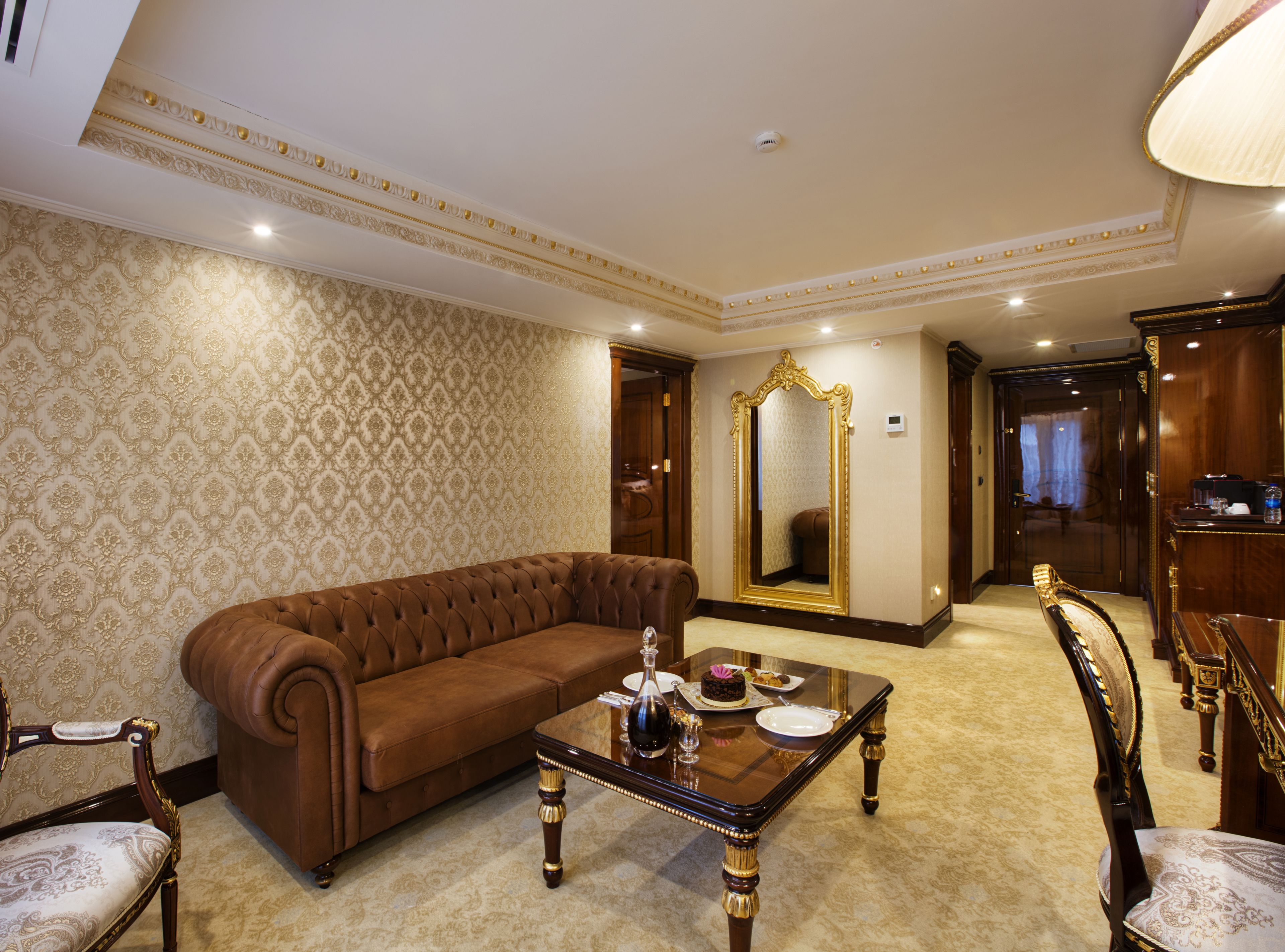 Une chambre luxueuse avec un canapé en cuir marron, une table basse en verre, des fauteuils décoratifs et un grand miroir.