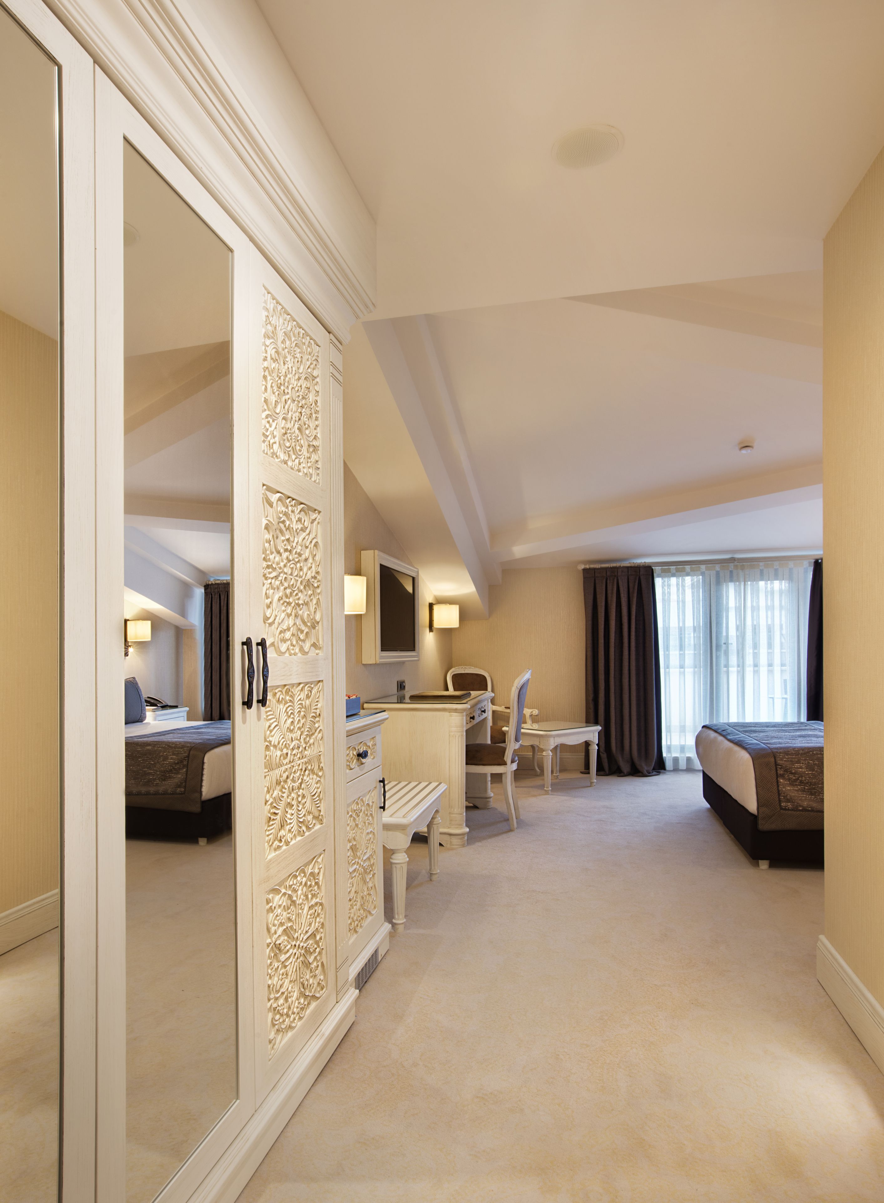 A hotel room with a large wardrobe, a desk with a chair, a bed, and a window with curtains.