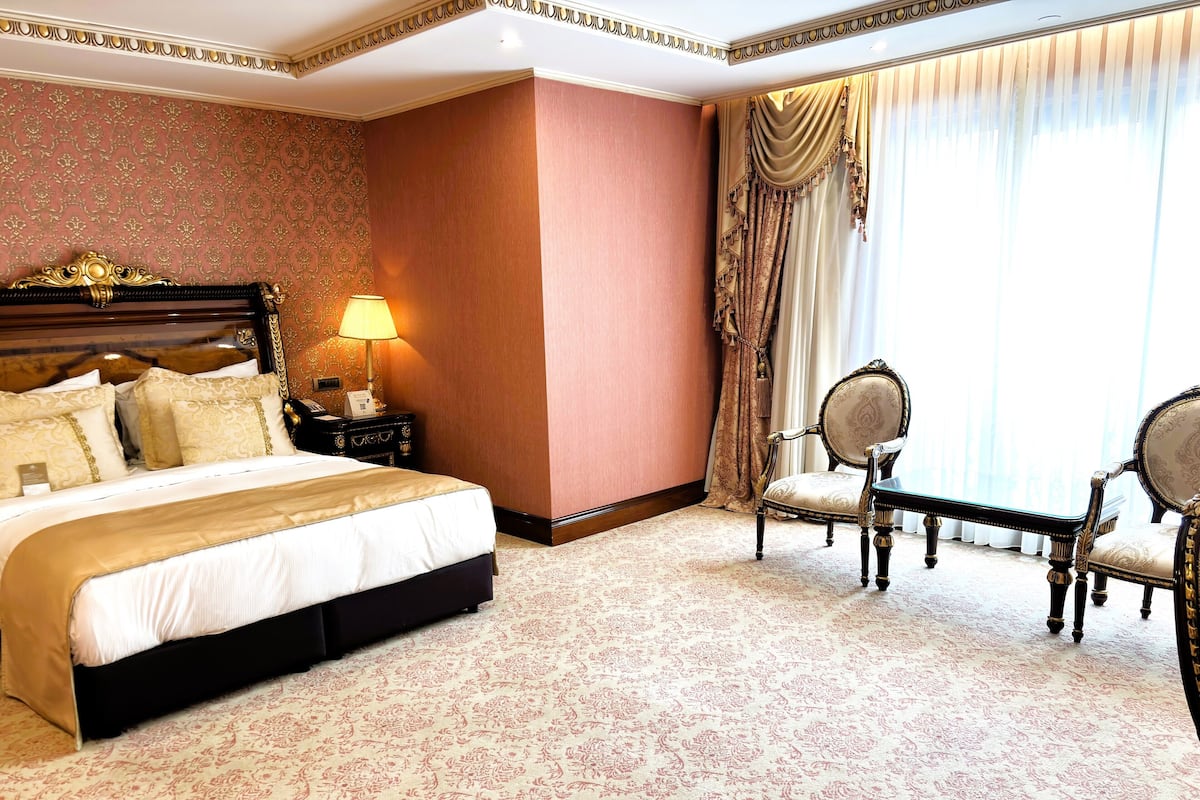 A hotel room with a large bed, a nightstand with a lamp, two upholstered chairs, and a view of the outdoors through sheer curtains.