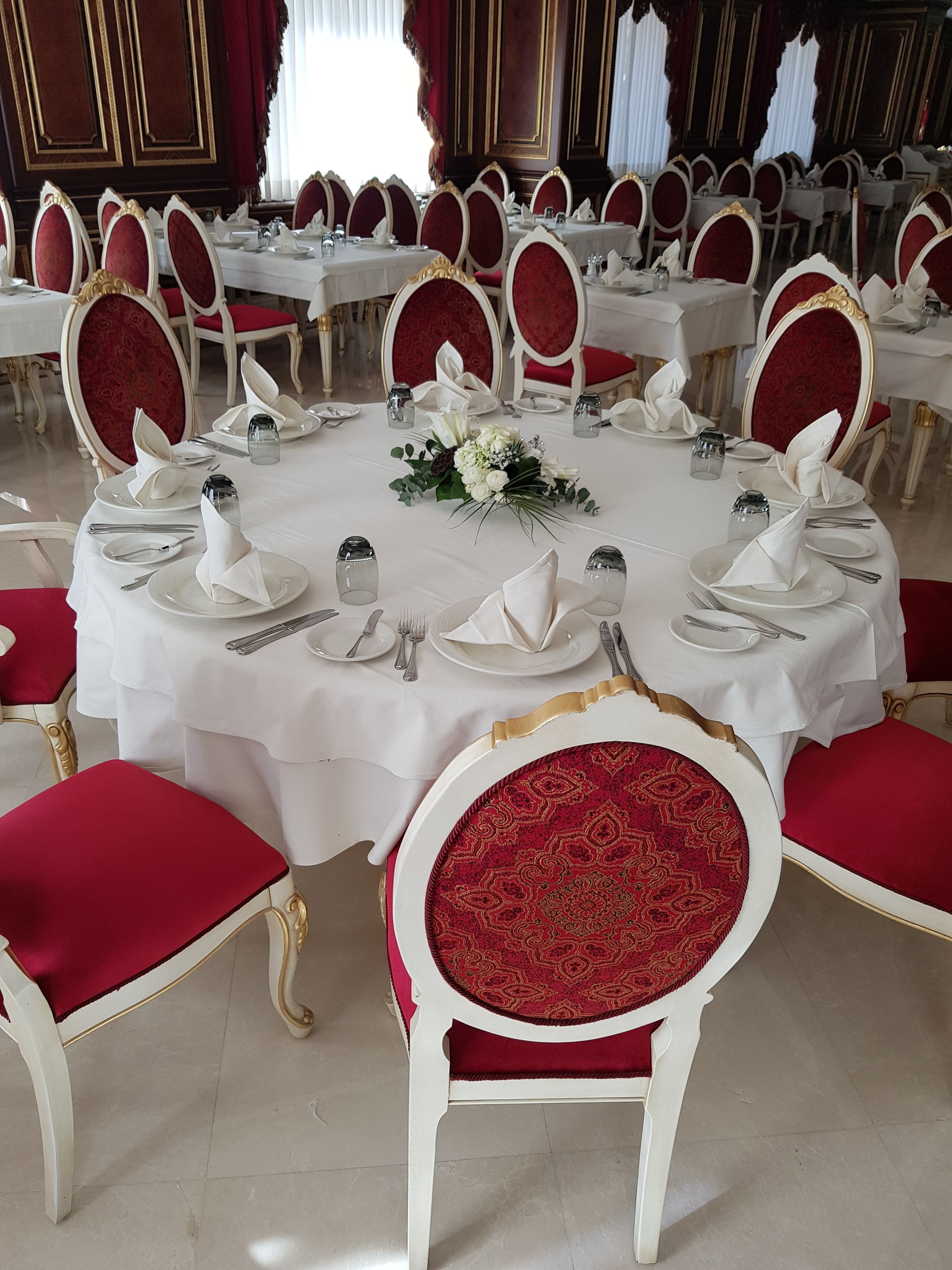 A banquet hall with round tables set for a formal event, each with white tablecloths, red upholstered chairs, and neatly arranged silverware and glassware.