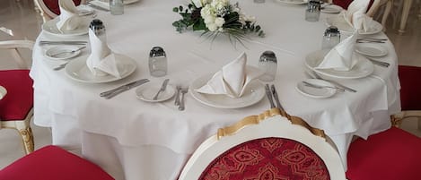 A banquet hall with round tables set for a formal event, each with white tablecloths, red upholstered chairs, and neatly arranged silverware and glassware.