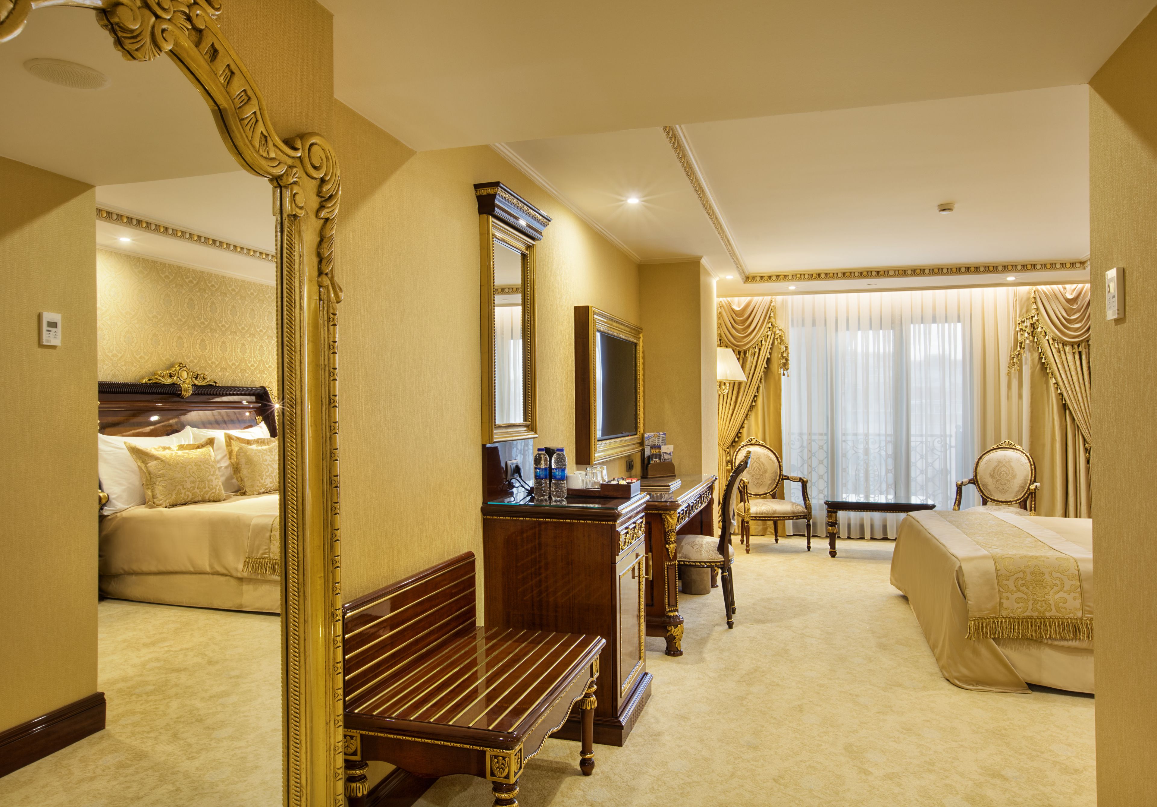 A hotel room with a large ornate mirror, a wooden bench, a desk with a computer, a chair, a bed, and a large window with curtains.