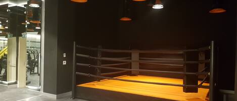 Un ring de boxeo con suelo de madera y cuerdas de metal, rodeado por una pared oscura con una puerta de vidrio en un lateral.