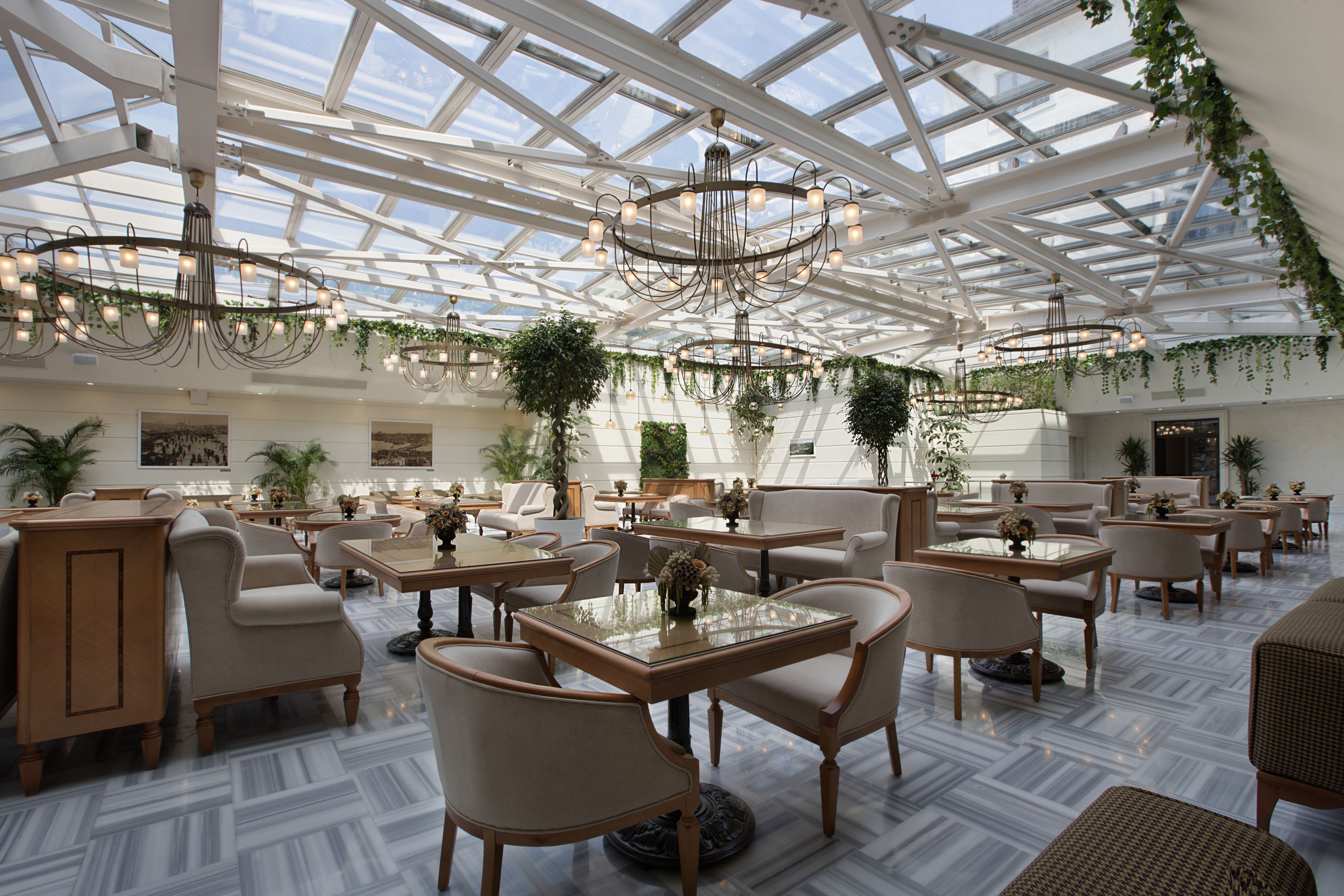 A spacious indoor area with multiple seating arrangements, including tables and chairs, under a large glass ceiling with natural light.