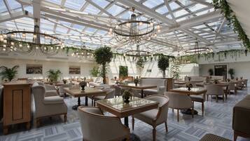 A spacious indoor area with multiple seating arrangements, including tables and chairs, under a large glass ceiling with natural light.