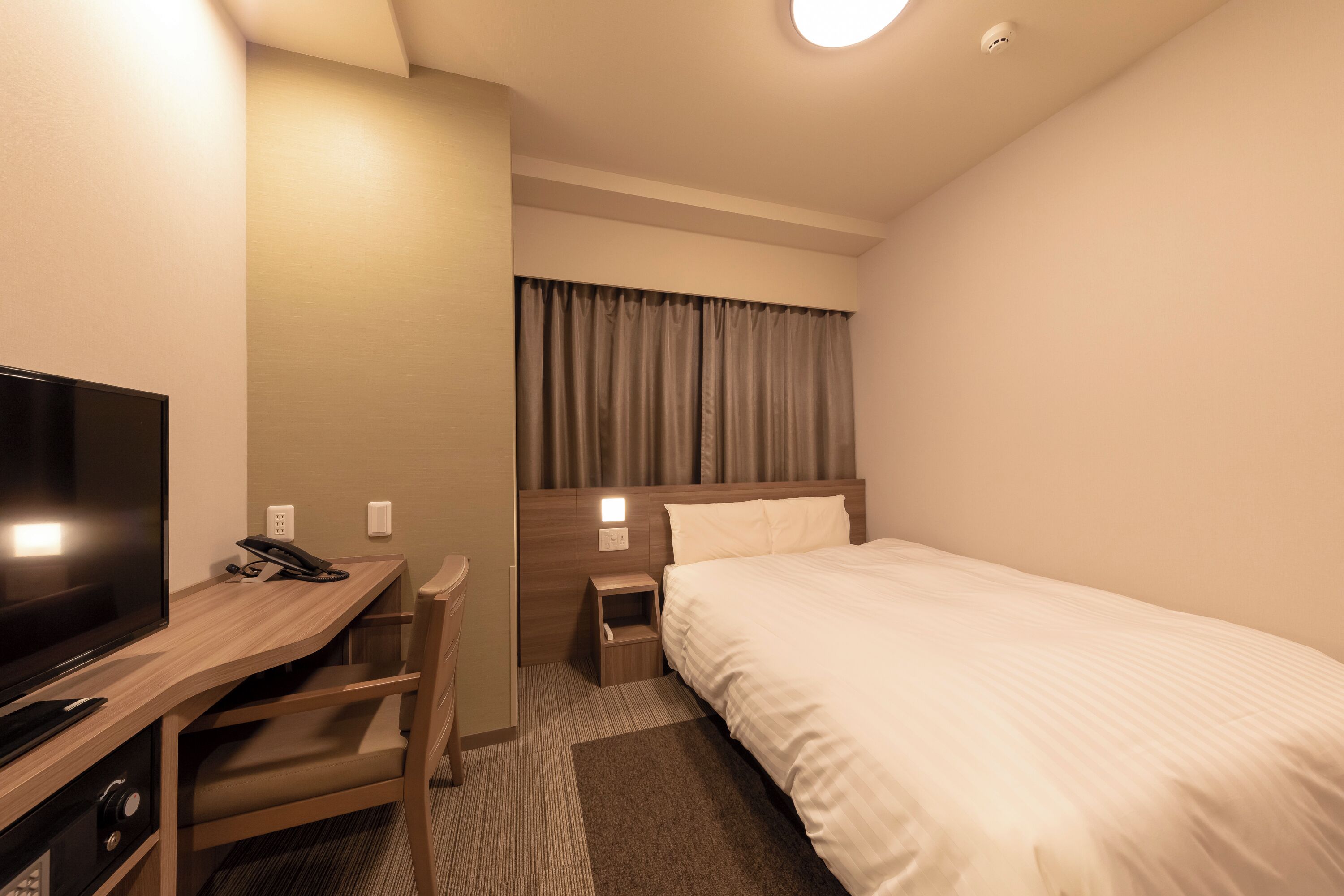 A hotel room with a bed, a desk with a television, a telephone, and a nightstand with a lamp.