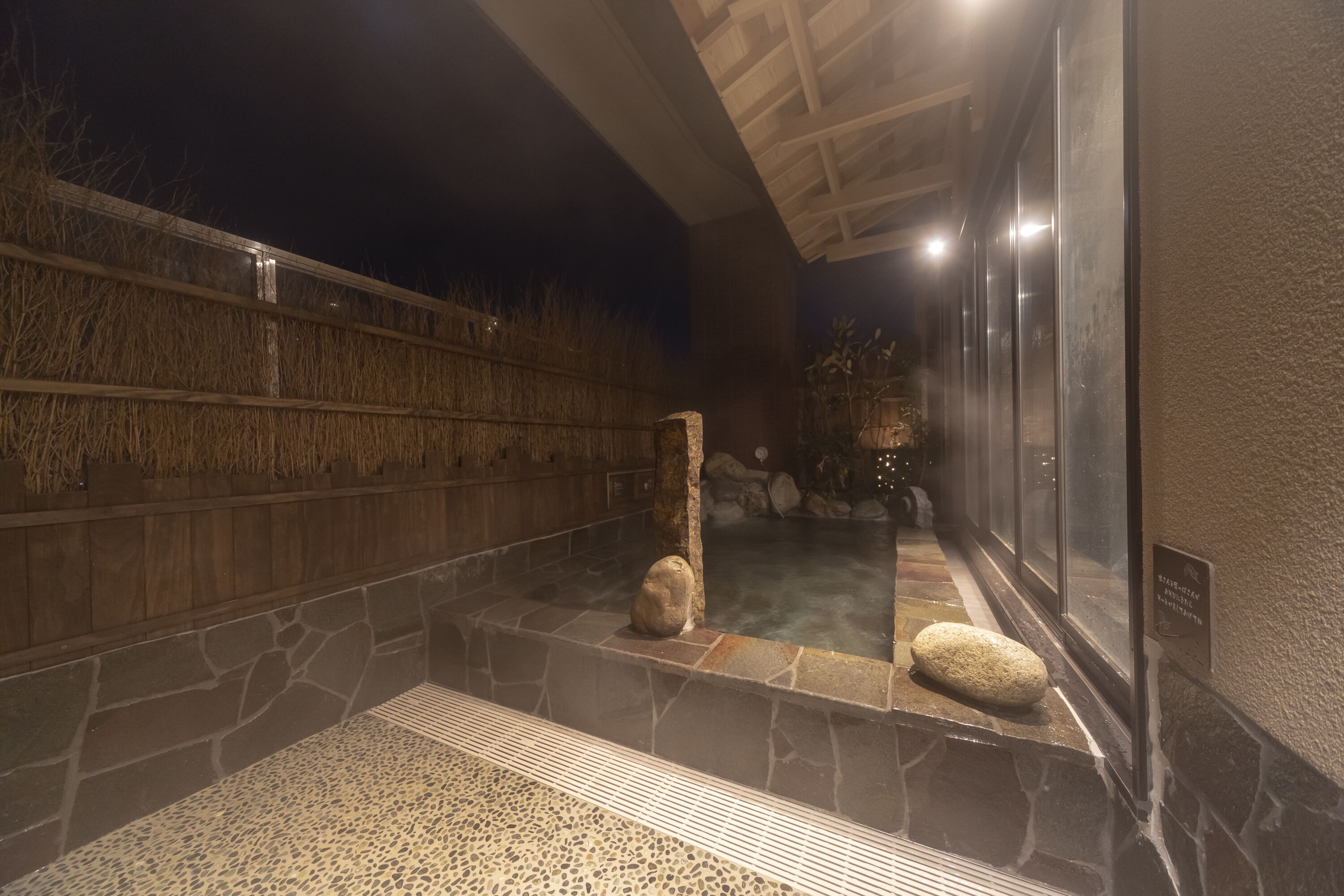 A stone-walled hot spring area with a wooden fence, a stone bench, and a glass enclosure.