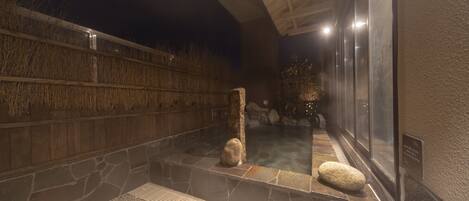 A stone-walled hot spring area with a wooden fence, a stone bench, and a glass enclosure.