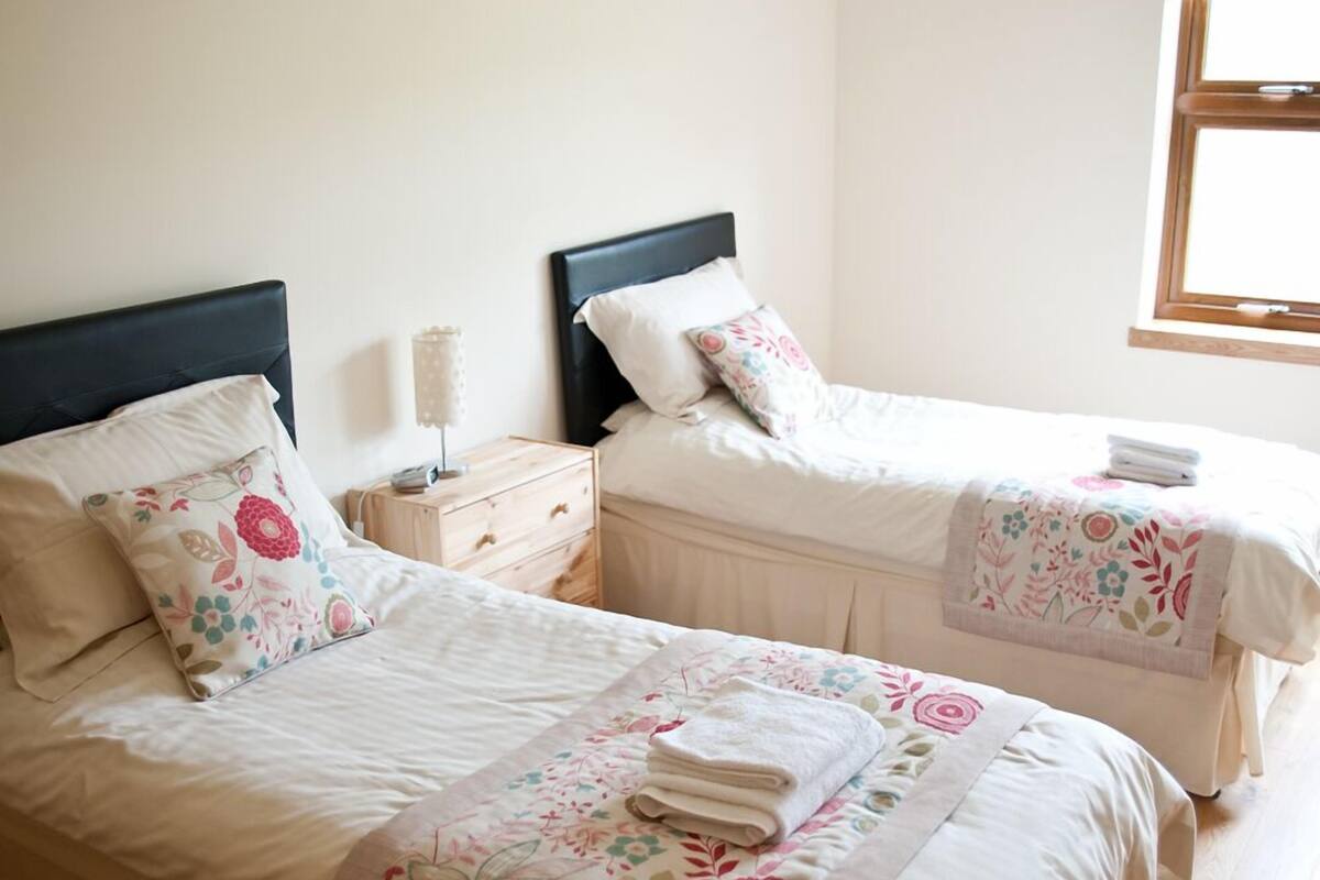 Quarto com duas camas, cada uma com roupa de cama e cabeceiras florais. Há uma mesa de cabeceira com abajur entre as camas.