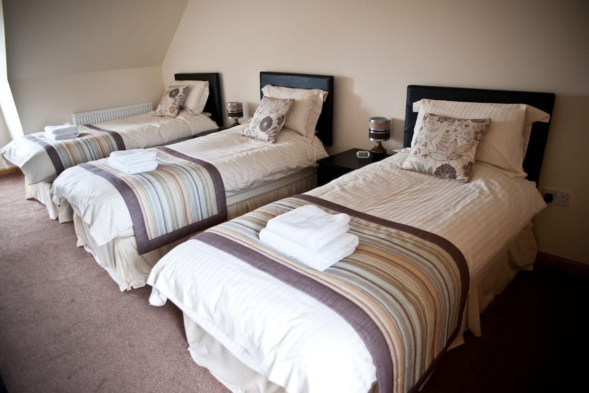 Quarto de hotel com três camas, cada uma com cabeceira, mesas de cabeceira e luminária.