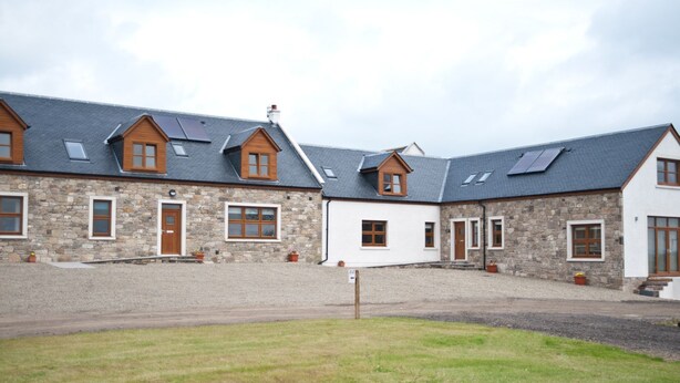 A row of stone and wooden buildings with multiple windows and doors, a gravel driveway, and a grassy area in the foreground.