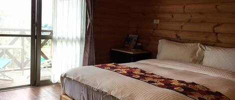 Un dormitorio de cabaña de madera con una cama grande, una mesita de noche y vistas al exterior a través de una puerta corrediza de vidrio.