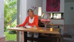 A woman sitting at a wooden table in a room with a large window, a glass fishbowl with pink flowers, and a red rose painting on the wall. - Hotel Rosenhaus Garni (Velbert)