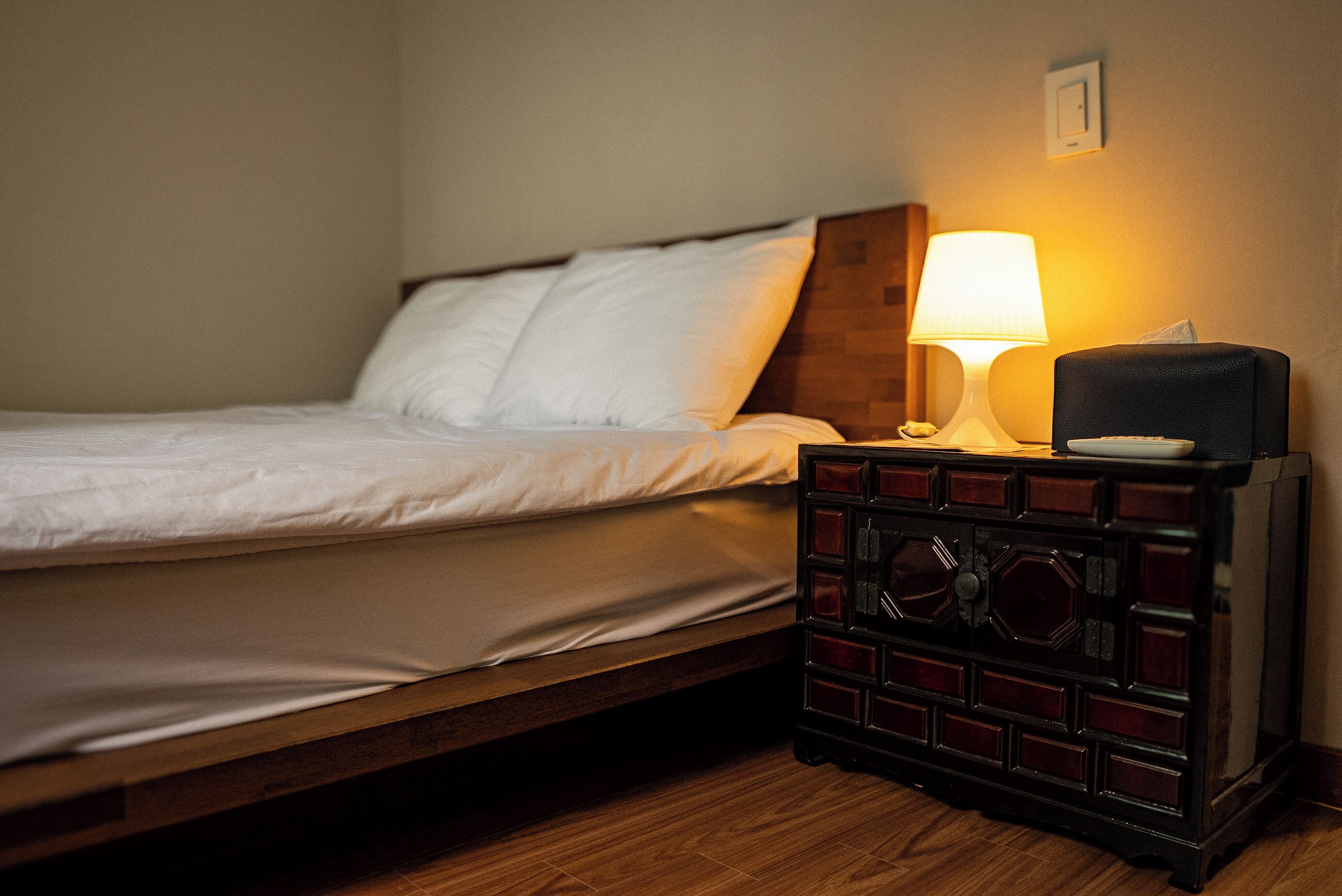 Una cama individual con cabecera de madera, una mesita de noche con lámpara y un mueble pequeño con una caja de pañuelos.
