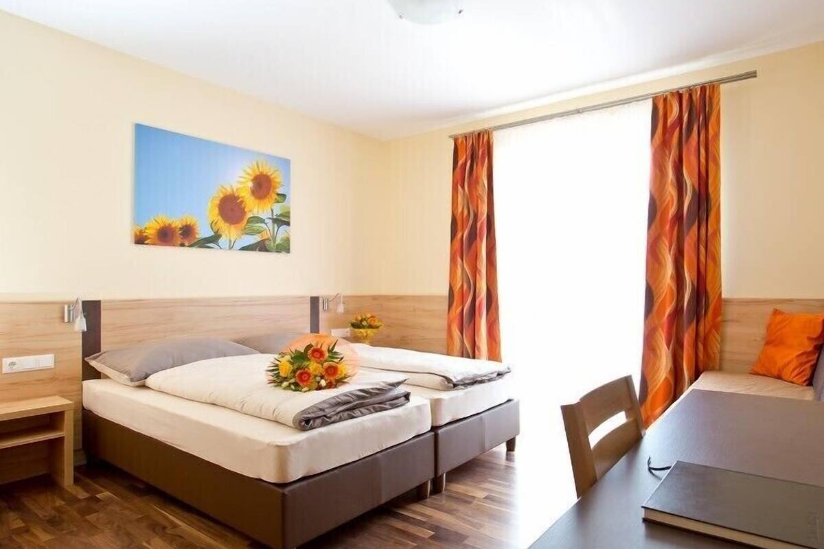 A hotel room with two beds, a desk, and a chair. There is a painting of sunflowers on the wall, and the room has a wooden floor.