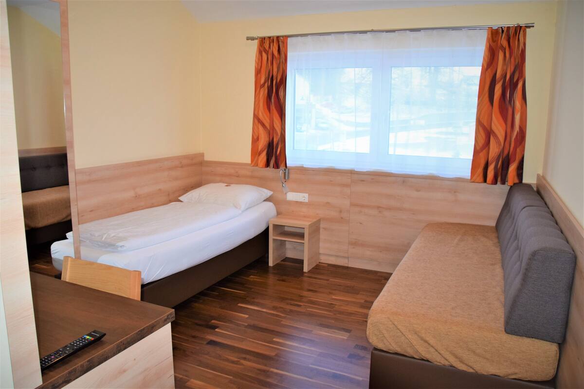 A small, compact room with a bed, a sofa, a small table, and a window with curtains.