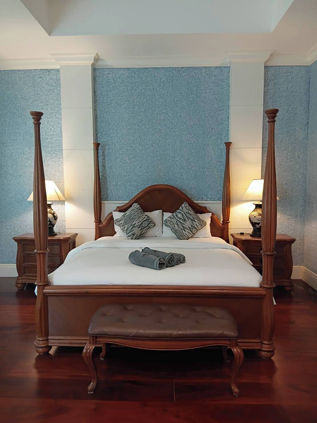 A four-poster bed with a wooden headboard, two nightstands with lamps, and a bench at the foot of the bed.