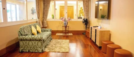 Una sala de estar con un sofá, una mesita con un florero, un ventilador y un televisor sobre un mueble bajo.