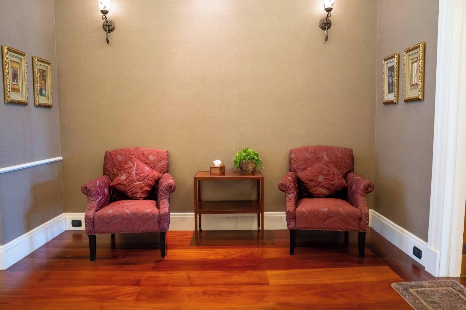 Une pièce avec deux fauteuils rouges, une petite table d’appoint en bois sur laquelle est posée une plante, et un mur beige orné de tableaux encadrés.
