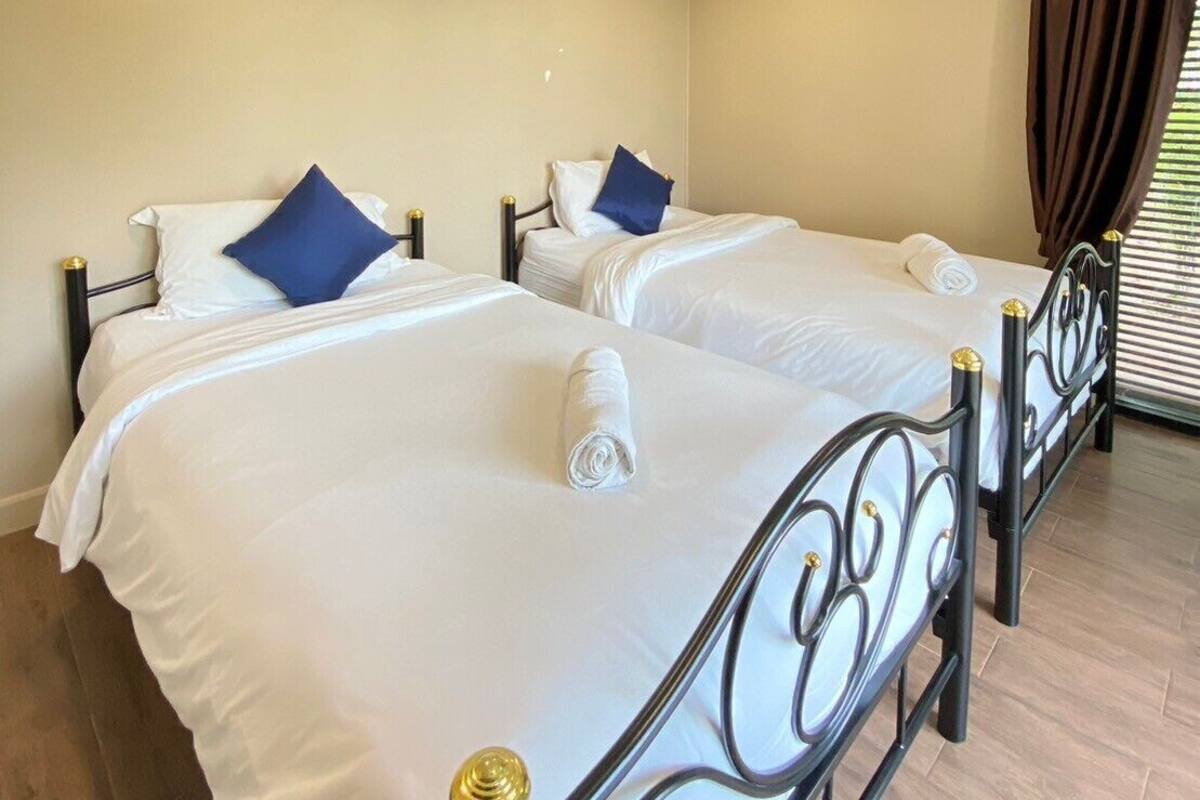 A hotel room with two beds, each with white bedding and blue pillows, a ceiling-mounted air conditioner, and a window with brown curtains.