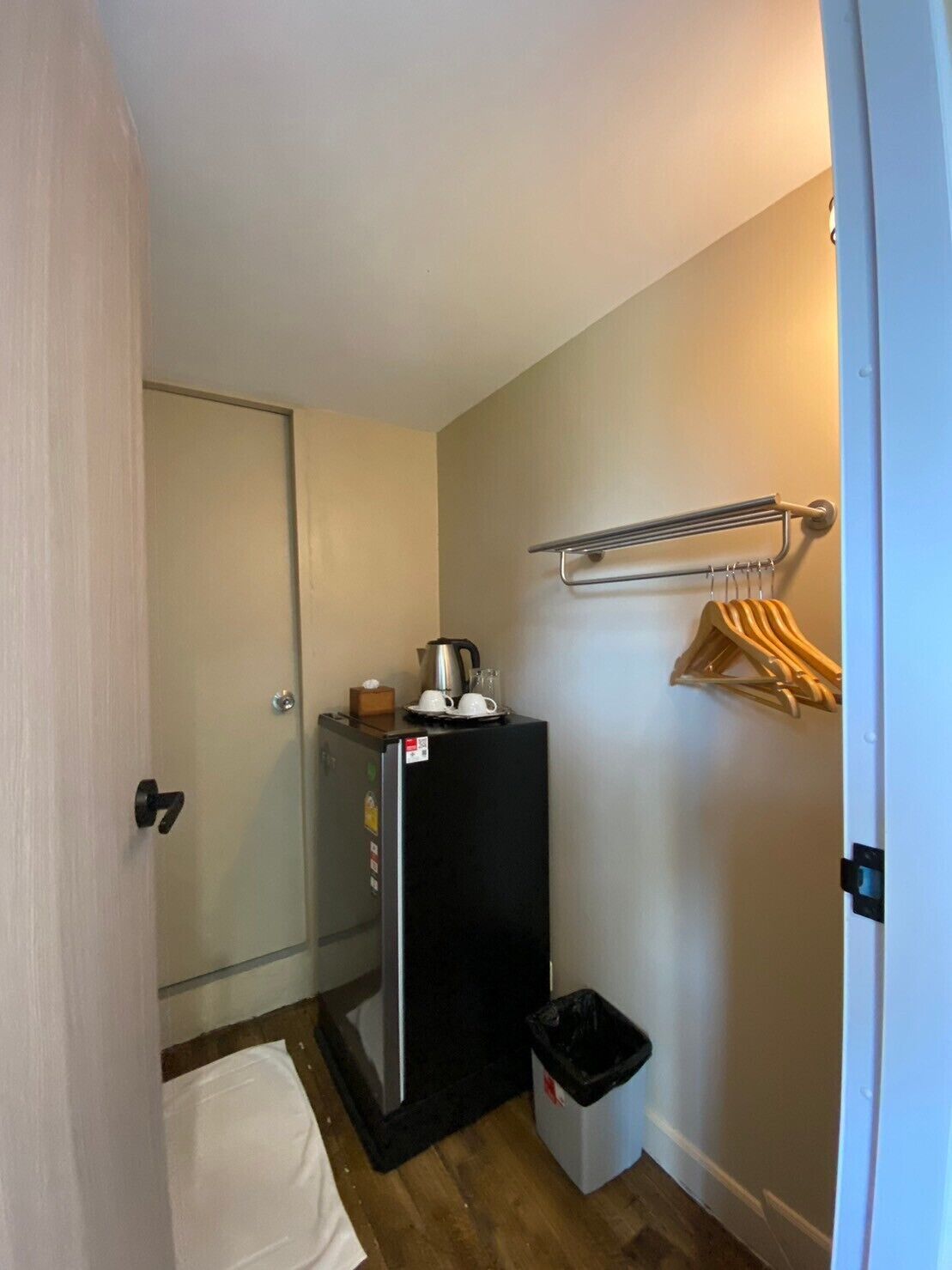 A compact bathroom with a toilet, a small sink, a black refrigerator, a trash bin, and a towel rack with a few towels.