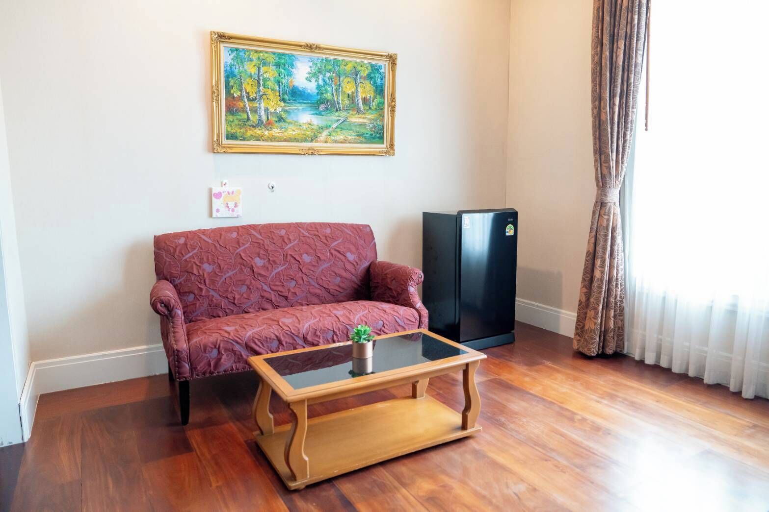 Un soggiorno con un divano rosso a fantasia, un tavolino da caffè in vetro con una pianta, un frigorifero nero e un quadro appeso al muro.