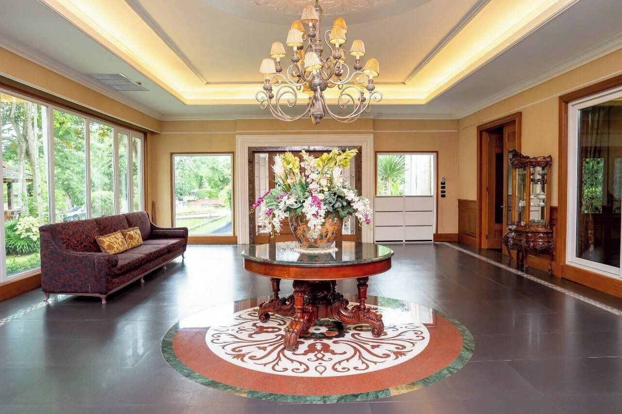 Eine großzügige Lobby mit einem großen Kronleuchter, einem runden Tisch mit floralem Arrangement, einer Couch und großen Fenstern mit Blick auf Grünflächen.