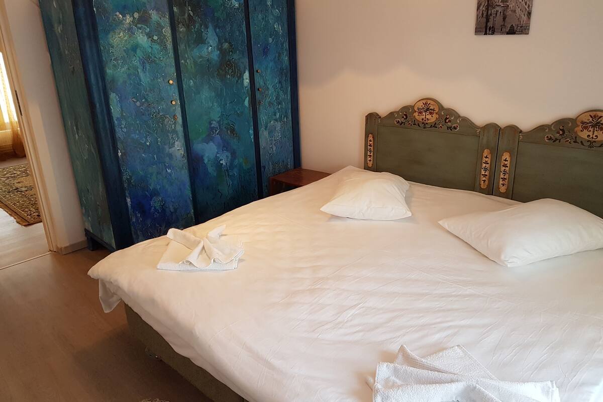 A neatly made bed with white linens and pillows, a wooden headboard with decorative carvings, and a blue wardrobe with a distressed finish.